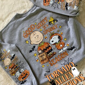May include: A heather gray sweatshirt featuring a Peanuts Halloween graphic. The design includes Charlie Brown and Snoopy in pumpkin costumes with the text "GOOD GRIEF IT'S SPOOKY SEASON" and "TRICK OR TREAT". The sleeves also display matching character prints.