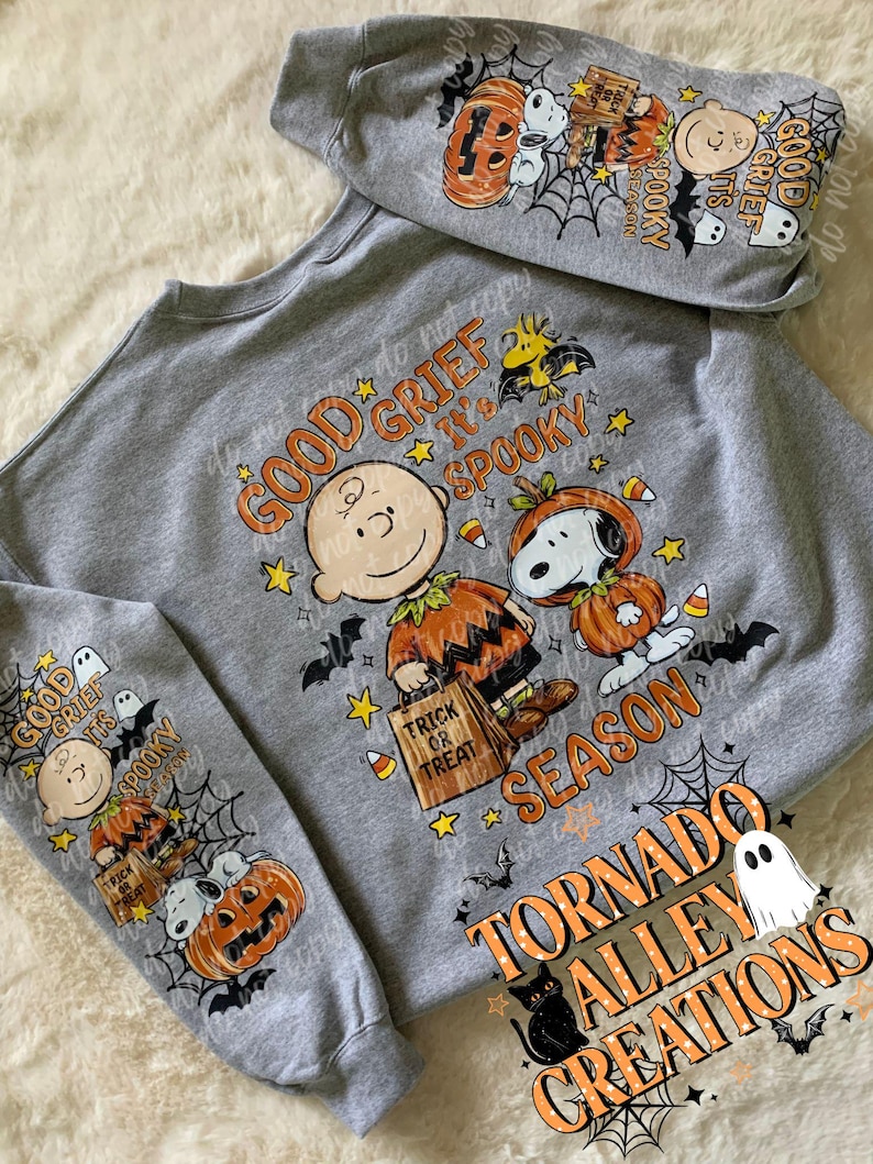 May include: A heather gray sweatshirt featuring a Peanuts Halloween graphic. The design includes Charlie Brown and Snoopy in pumpkin costumes with the text "GOOD GRIEF IT'S SPOOKY SEASON" and "TRICK OR TREAT". The sleeves also display matching character prints.