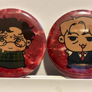 May include: Two circular pin-back badges featuring chibi-style character illustrations against a red, marbled meat-like background. The left badge shows a person with dark hair and glasses in a green shirt. The right badge shows a blonde person in a suit holding a small knife.