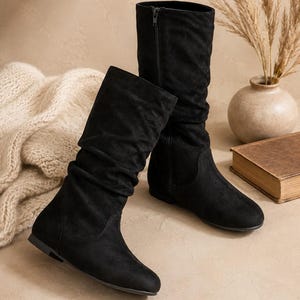 May include: A pair of black mid-calf boots featuring a soft, slouchy shaft design, a rounded toe, and a flat sole. Each boot includes a vertical side zipper closure and a matte finish, providing a simple and functional footwear option.