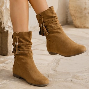 Puede incluir: Botas de media caña en ante color canela con diseño de caña holgada, punta redondeada, suela plana y correas decorativas envolventes adornadas con borlas a juego. Las botas tienen una textura mate suave y un estilo sencillo de poner.