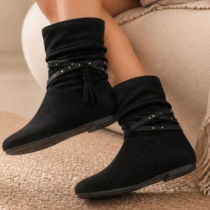 May include: A pair of black mid-calf boots featuring a soft, slouchy upper, a rounded toe, and a flat sole. Each boot is accented with a thin, crisscrossing strap detail adorned with small silver-toned studs and a decorative side tassel.