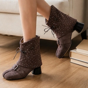 May include: A pair of dark brown ankle boots featuring a fold-over cuff with a subtle white speckled pattern, a thin lace-up closure at the front, a decorative buckle strap across the vamp, and a sturdy black cylindrical block heel.