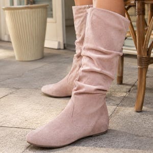 May include: A pair of knee-high boots in a soft, dusty pink color. These boots feature a slouchy, gathered shaft design, a rounded toe, and a flat sole. The exterior has a matte, suede-like texture, and a vertical zipper is visible on the inner side.