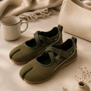 May include: A pair of olive green split-toe shoes featuring a textured suede-like finish, crisscross straps with hook-and-loop closures, a pull tab at the heel, and a flat rubber sole. The design includes a distinct separation between the big toe and the other toes.