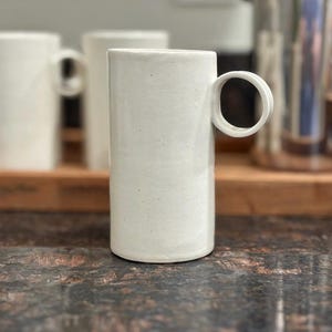Puede incluir: Una taza de cerámica blanca alta y cilíndrica con un acabado mate suave y un asa circular distintiva unida al lateral. La taza presenta un diseño vertical limpio con una base plana y una superficie exterior sencilla y sin adornos.