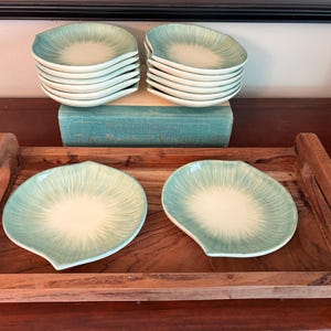 May include: A set of small, leaf-shaped ceramic dishes featuring a white center that fades into a light teal, radiating edge. Two dishes are placed on a rectangular wooden tray, while two stacks of five dishes rest on a teal-colored book.
