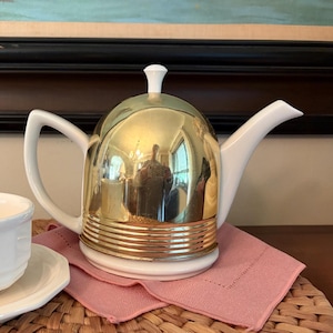 May include: A teapot featuring a white ceramic base, handle, and spout, paired with a polished gold-tone metal dome cover. The dome has a smooth upper section and a ribbed lower band. A small white knob sits atop the center of the gold dome.