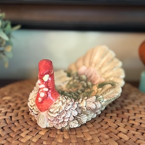 May include: A ceramic turkey-shaped serving dish featuring a bright red head and neck with a textured, cream-colored body and tail feathers. The piece has a hollow center with a detailed, molded feather design across the back and wings.
