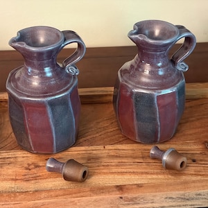 May include: Two ceramic cruets with a glossy purple and dark grey striped glaze, each featuring a curved handle and a pouring spout. Two separate tapered stoppers with a brown finish are placed in front of the vessels on the wooden surface.