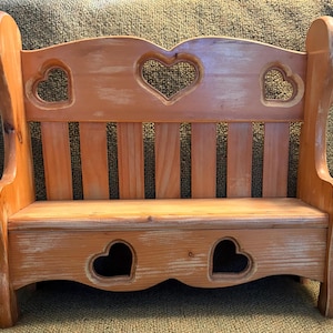 May include: A light brown wooden bench featuring a high backrest with three heart-shaped cutouts and a seat base with two additional heart-shaped cutouts. The wood grain is visible throughout the structure, which has a smooth, polished finish.