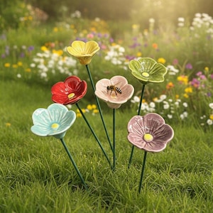 Puede incluir: Un conjunto de seis estacas de jardín con flores de cerámica en amarillo, rojo, azul claro, rosa, verde y morado, cada una con un centro amarillo. Una pequeña abeja está posada sobre la flor rosa. Cada flor está unida a un tallo de metal verde.