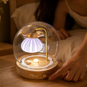 May include: A round glass dome lamp with a wooden base featuring the name Olivia. Inside the dome, a gold-toned curved arm supports a glowing purple sea urchin shell topped with a starfish, with small shells resting on the wooden base.