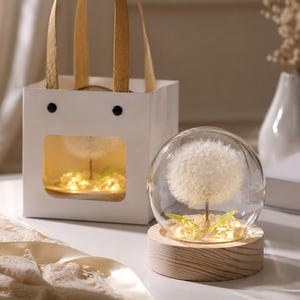 May include: A white gift bag with two handles and a square cutout window containing a dandelion inside a glass dome on a wooden base. Beside it sits a matching dandelion inside a glass dome on a light wood base, illuminated by small warm LED lights.