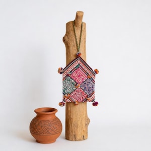 May include: A small, rectangular, fabric wall hanging featuring a geometric pattern in red, green, and blue, accented with colorful pom-poms along the edges. It hangs from a green cord. Beside it sits a small, round, terracotta-colored clay pot.