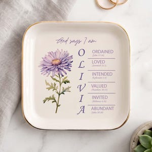 May include: A square white ceramic trinket dish with gold trim featuring a purple chrysanthemum illustration and the text God says I am OLIVIA followed by the words ORDAINED, LOVED, INTENDED, VALUED, INVITED, and ABUNDANT with corresponding Bible verses.
