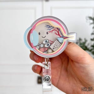 Op de afbeelding: Een ronde acryl badgehouder met een dwarsdoorsnede van een menselijk oog met een lachend gezicht. In de doorzichtige behuizing zitten kleine zwevende bedeltjes, waaronder een paar ogen, een roze flesje en een witte oogtestkaart met letters.