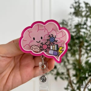 Op de afbeelding: Een roze badgehouder in de vorm van een brein met een lachend gezicht. Het ontwerp bevat een klein breinicoon, twee gestileerde neuronen, een koffiebeker, een paarse pil en een gele bliksemschicht. De badgehouder heeft een doorzichtige clip.