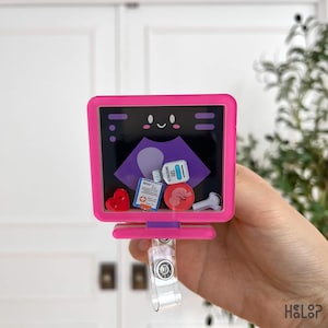 May include: A pink rectangular badge reel featuring a screen display with a smiling face and an ultrasound image. Inside the frame are small charms including a red heart, a medical chart, a bottle of ultrasound gel, a fetus, and a probe.