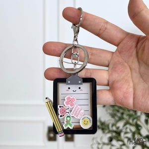May include: A keyring featuring a black-framed clipboard charm with a white paper insert. The clipboard displays a smiling face, a yellow pencil, and small charms including a pink bandage, a brain, a green ribbon, and a white circle with the text Trust Yourself and Mental Health Matters.