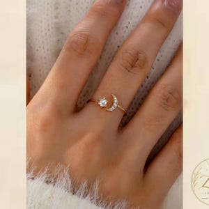 Könnte beinhalten: Ein goldfarbener Ring mit einer zarten Mondsichelform, die mit kleinen klaren Steinen und einem einzelnen runden, sternförmig gefassten klaren Stein verziert ist, was ein himmlisches Design erzeugt. Der Ring ist dünn und poliert.