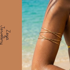 Peut inclure: Un bracelet de bras en métal doré au design multicouche enveloppant qui entoure le haut du bras. La pièce est présentée avec le texte Zeya Jewelery sur un fond beige uni, avec une scène de plage tropicale floue en arrière-plan.