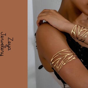 Könnte beinhalten: Ein goldfarbenes Armband und ein Oberarmreif, beide mit einem filigranen, durchbrochenen zweigartigen Muster. Der Text Zeya Jewelery ist vertikal auf der linken Seite des Bildes in einer schwarzen, kursiven Schriftart dargestellt.