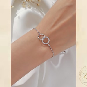Puede incluir: Una pulsera de cadena en tono plateado con dos dijes circulares entrelazados. Los círculos centrales están adornados con pequeñas piedras claras y brillantes engastadas a lo largo de los bordes exteriores. La pulsera tiene un diseño de cadena fina.