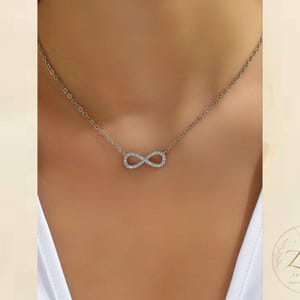 May include: A silver-toned necklace featuring a central infinity symbol pendant encrusted with small, clear, sparkling stones. The pendant is attached to a delicate, thin chain that rests around the neck. The logo Zeya Jewelry is visible.