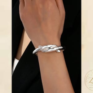 Peut inclure: Un bracelet manchette en métal argenté poli présentant un design de nœud lisse et entrelacé. La surface métallique est réfléchissante et incurvée, formant une boucle continue qui entoure le poignet. Le logo Zeya Jewelry est visible.