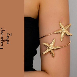 Op de afbeelding: Een goudkleurige metalen armband voor de bovenarm met twee gestructureerde zeester-motieven. Het sieraad heeft een dunne, flexibele band die om de arm wikkelt. De tekst Zeya Jewelery staat in het zwart aan de linkerkant van de afbeelding.