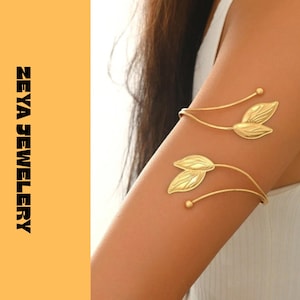 Peut inclure: Un bracelet de bras en métal doré composé de deux tiges incurvées se terminant chacune par une paire de feuilles texturées et une petite perle sphérique. Le texte ZEYA JEWELERY est imprimé verticalement en noir sur le côté gauche de l'image.