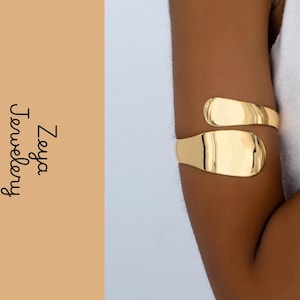 May include: A polished gold-tone arm cuff featuring a smooth, open-ended wrap design with rounded, flattened ends. The text Zeya Jewelery is printed in black on the left side of the image against a solid tan background.
