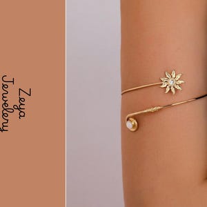 Puede incluir: Un brazalete dorado con un dije en forma de sol con una piedra central transparente y un extremo con una piedra blanca redonda. El texto Zeya Jewelery es visible en el lado izquierdo de la imagen sobre un fondo de color beige.