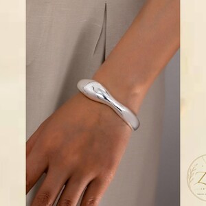 Puede incluir: Un brazalete de tono plateado pulido con una forma suave, orgánica y fluida. La pulsera presenta un diseño grueso y redondeado que se estrecha ligeramente, creando una superficie continua y reflectante alrededor de la muñeca.