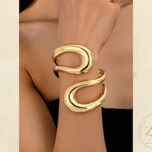 Puede incluir: Un brazalete de tono dorado pulido con un diseño abierto, fluido y asimétrico de doble bucle que se envuelve alrededor de la muñeca. La superficie es lisa y reflectante, creando una forma continua e interconectada con un acabado de alto brillo.