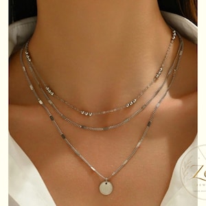 Può includere: Una collana a strati color argento composta da tre catene distinte. La catena superiore presenta piccole perline metalliche, la catena centrale ha maglie piatte rettangolari e la catena inferiore presenta un piccolo ciondolo a disco rotondo.