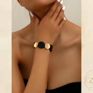 Puede incluir: Una pulsera que presenta elementos circulares alternos en metal dorado pulido y material negro mate. El diseño consiste en una secuencia de discos redondos unidos entre sí para formar una banda flexible que rodea la zona de la muñeca.