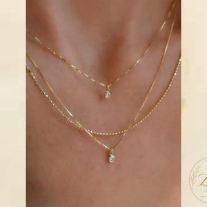 Puede incluir: Un conjunto de collares dorados en capas que consta de tres cadenas delicadas. La cadena superior presenta un pequeño colgante de piedra redonda transparente, mientras que la cadena inferior incluye una gota de piedra transparente.