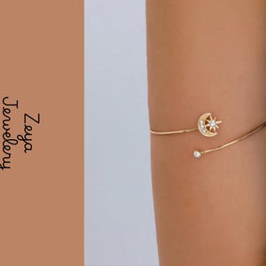 May include: A gold-toned arm cuff featuring a crescent moon and star motif adorned with small clear stones. One end of the open band terminates in a single clear stone. The text Zeya Jewellery is visible on the left side of the image.