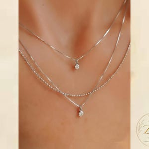 Puede incluir: Un conjunto de tres collares superpuestos en tono plateado. La cadena superior presenta un pequeño colgante de piedra redonda transparente, la cadena central tiene una textura de cuentas y la cadena inferior sostiene un colgante de piedra mayor.