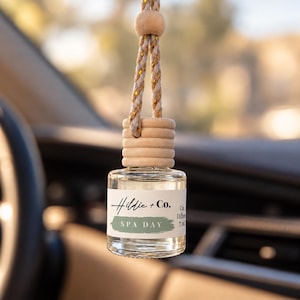May include: A clear glass car diffuser bottle with a light wood cap and a braided cord. The white label features the black text Hildie + Co. and the green text SPA DAY, with the additional text Car Diffuser 7 ml printed on the side.