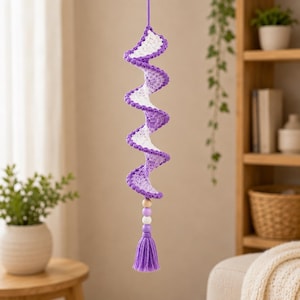 May include: A hanging crochet spiral ornament featuring alternating white and purple yarn sections. The spiral structure is suspended from a matching purple cord and finished at the bottom with a series of three wooden and plastic beads and a purple tassel.