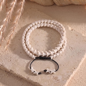 Könnte beinhalten: Ein zweireihiges Armband aus cremefarbenen, segmentierten, wirbelartigen Perlen. Die Stränge sind durch eine schwarze Kordel mit verstellbarem Schiebeknoten verbunden, an deren Enden sich zwei kleine, passende knochenartige Perlen befinden.