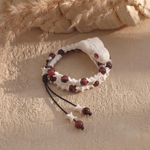 Könnte beinhalten: Ein Armband mit einem geschnitzten weißen Schlangenkopf mit roten Augen, abwechselnd mit dunkelroten runden Perlen und weißen knochenförmigen Zwischenstücken. Das Design umfasst silberfarbene Metallelemente und zwei schwarze Kordeln mit Perlen.
