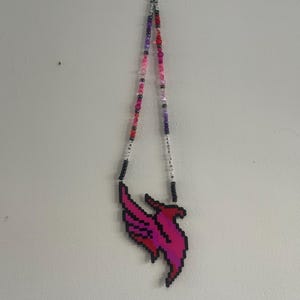 May include: A beaded necklace featuring a pixelated phoenix pendant crafted from small black, pink, and magenta beads. The necklace strand consists of various shades of pink, purple, and clear beads, with white letter beads spelling out words.