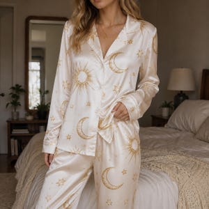 May include: A cream-colored satin pajama set featuring a long-sleeved button-down shirt with a notched collar and matching trousers. The fabric is decorated with a repeating gold celestial print consisting of suns, crescent moons, and stars.
