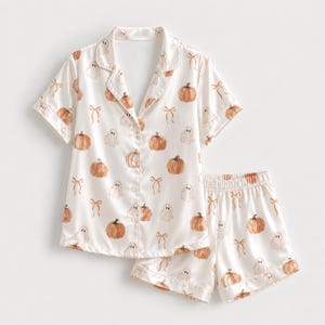 Op de afbeelding: Een tweedelige pyjamaset bestaande uit een shirt met korte mouwen en knoopsluiting en een bijpassende short. De witte stof heeft een herhalend patroon van oranje pompoenen, witte spoken en lichtbruine strikken, afgewerkt met een reverskraag.