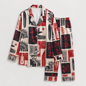 May include: A two-piece pajama set featuring a long-sleeve button-down shirt and matching pants. The fabric is cream-colored with a repeating block print pattern in red and black, depicting surfboards, tropical flowers, palm leaves, and ukuleles.