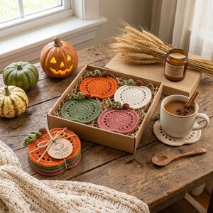 May include: A set of four round crochet pumpkin coasters in orange, green, white, and mauve, presented in a cardboard box. A matching stack of coasters is tied with twine and a tag labeled Fall Harvest Coasters, alongside a mug and pumpkins.